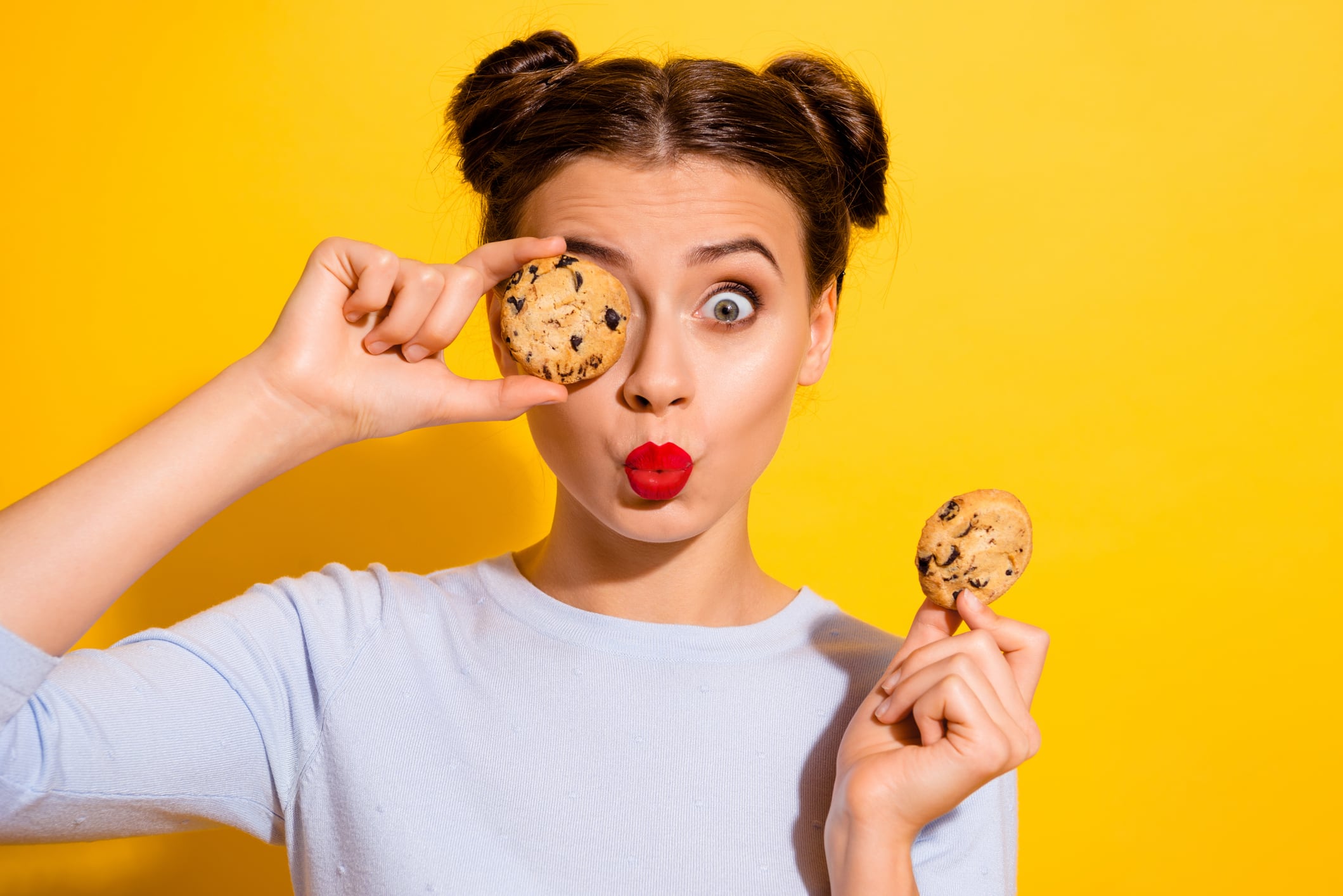 Woman wearing red lipstick and space buns in her hair, holding biscuit over one eye. Isolated yellow coloured background.