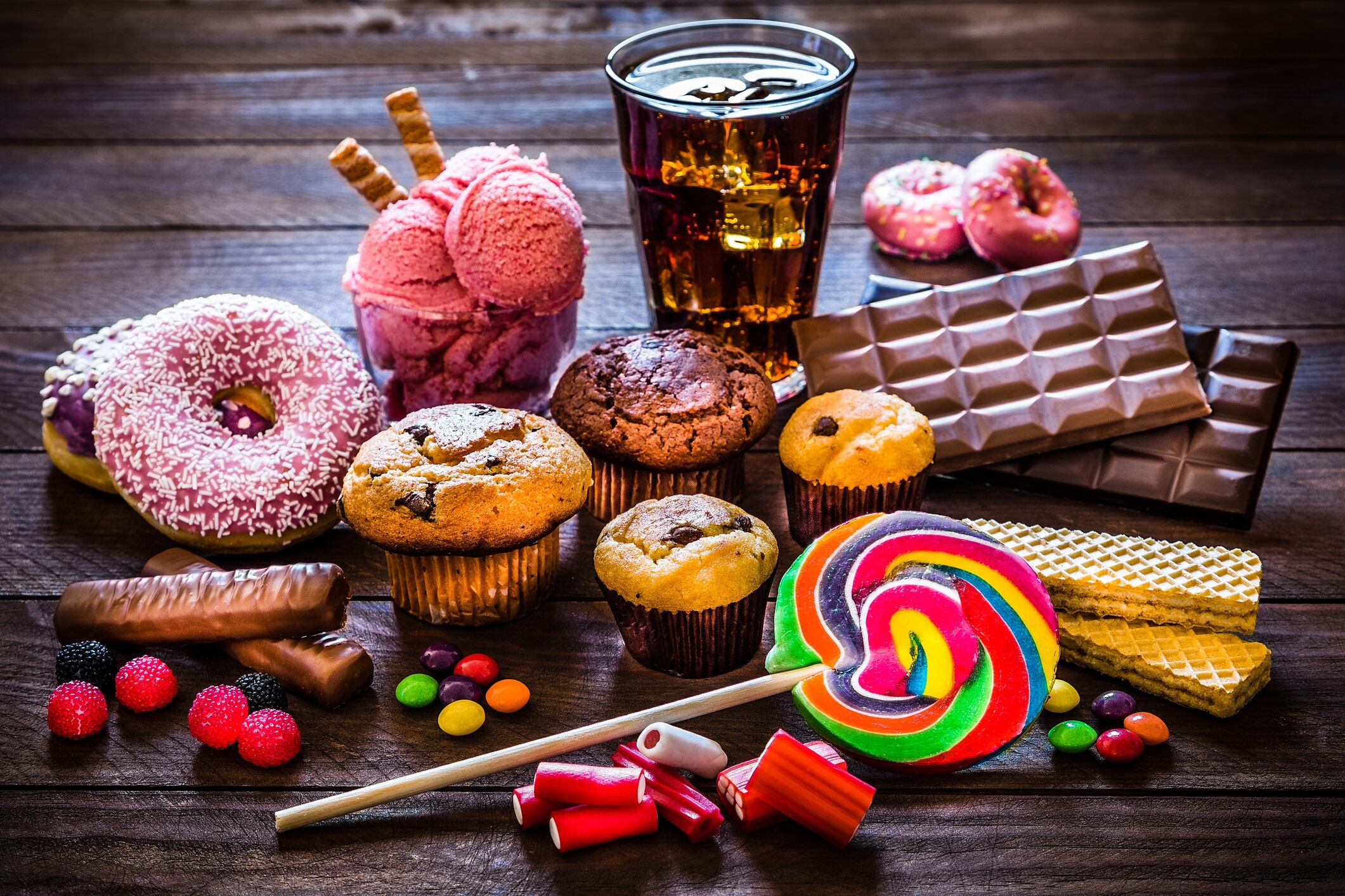 Assortment of products with high sugar level like candies, gummy candies, donuts, soda, chocolate, lollipop, wafers and cupcakes on rustic wooden table.