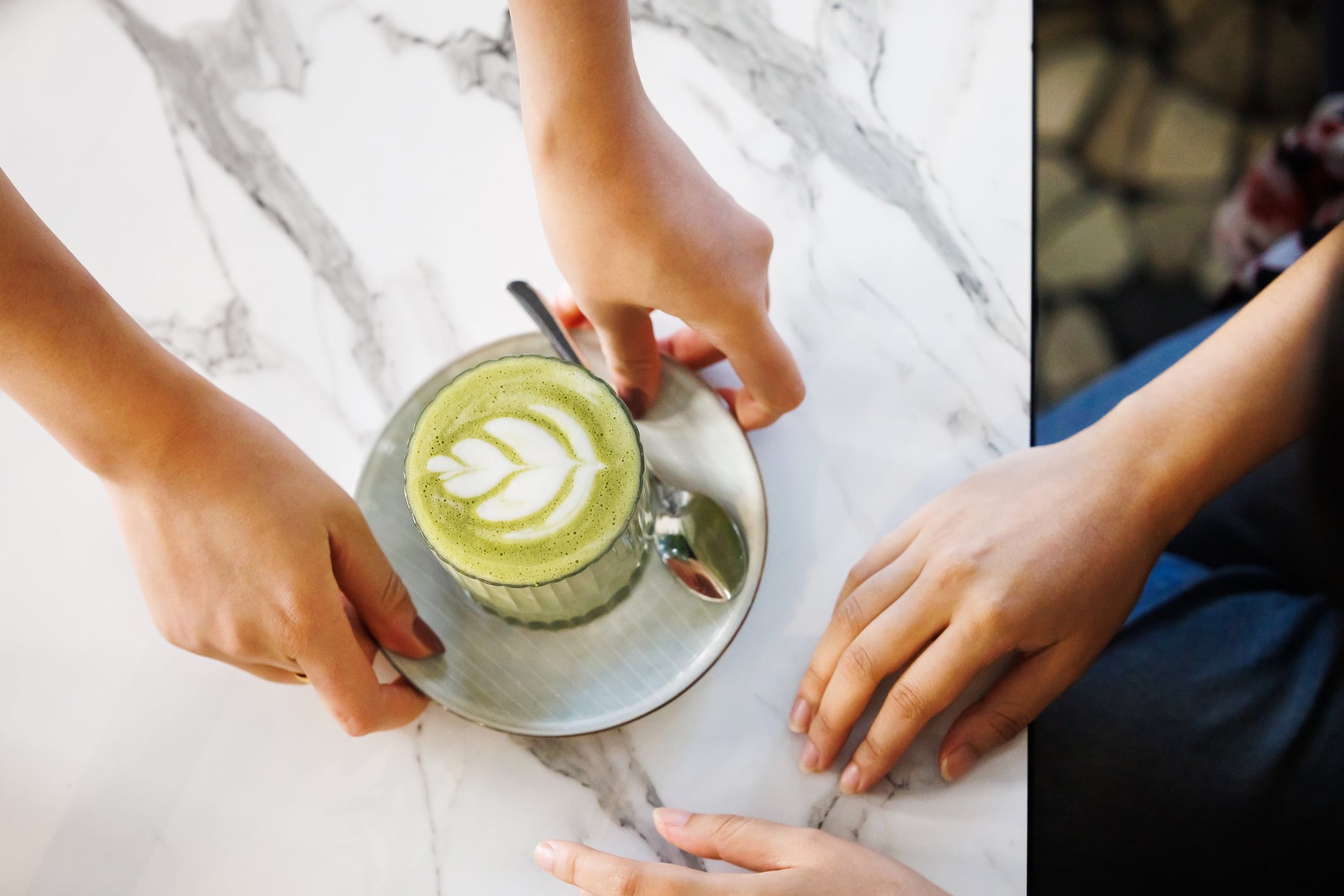 Overhead view of two hands reaching for a cup of matcha latte with artistic foam, placed on a marble surface.