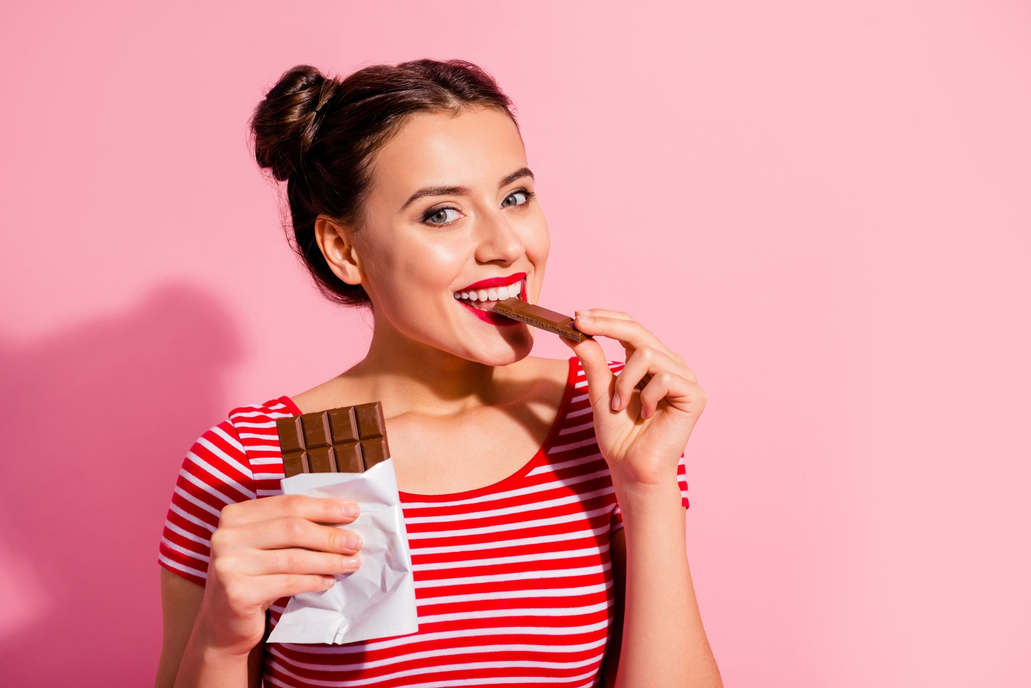 Woman in striped t-shirt eating a bar of chocolate. Pink pastel background.