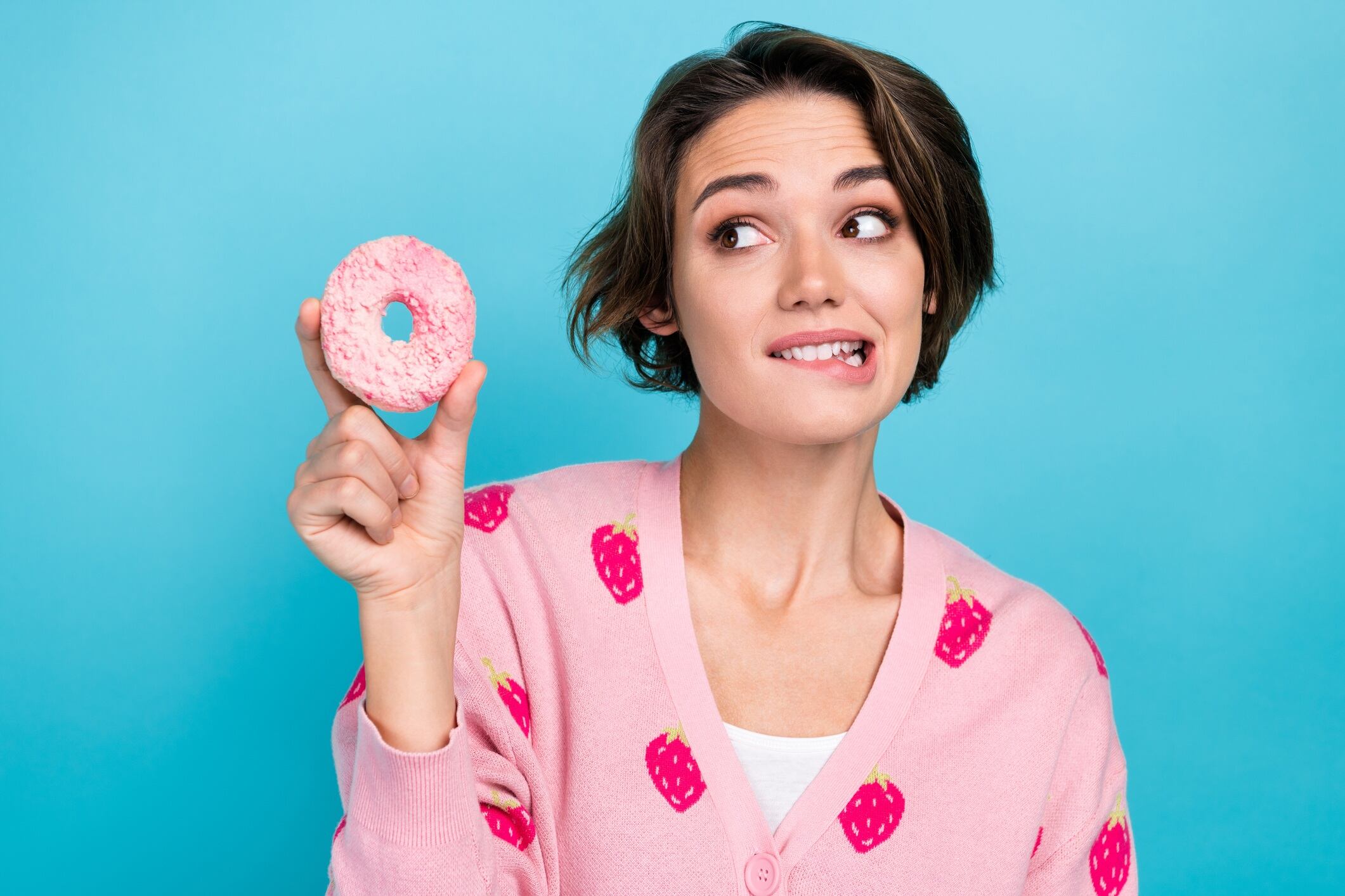 Woman holding doughnut and wearing strawberry-print cardigan. Blue background.
