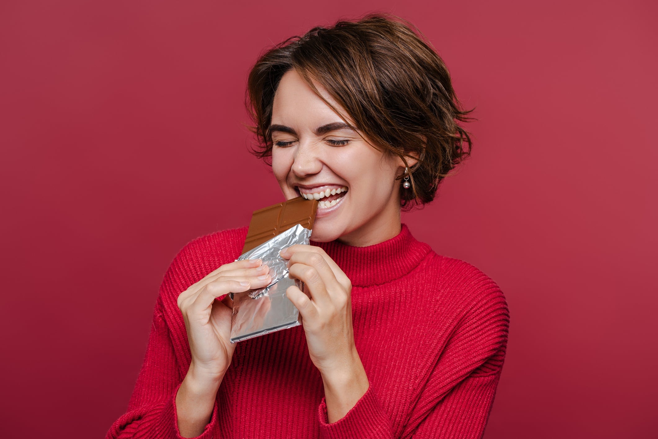 Woman wearing red jumper and eating chocolate bar - red background.