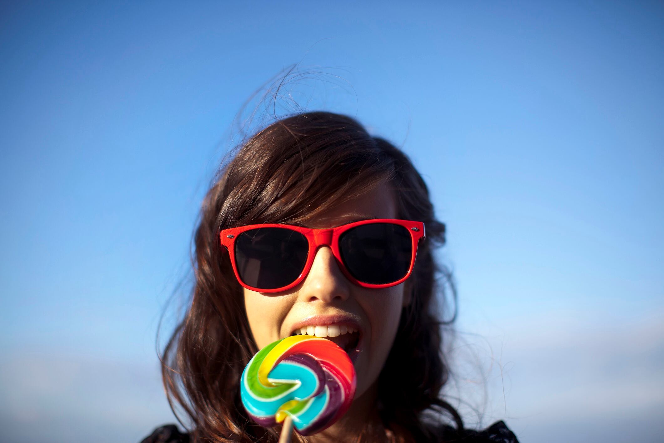 Young woman wearing red sunglasses eating a colourful lollipop.