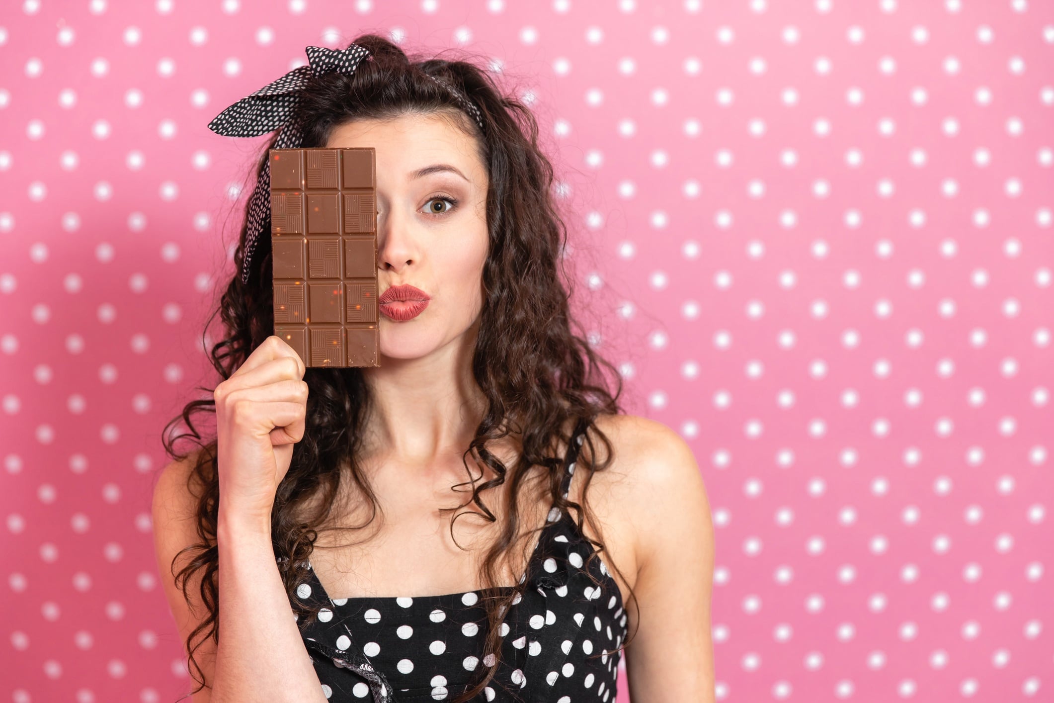 Woman hiding half of her fact behind a bar of chocolate. She's wearing a polka-dot dress and is standing in front of a polka-dot background.