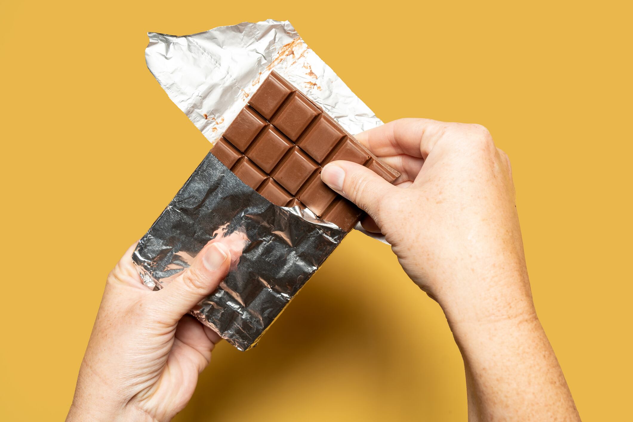 Close up of female hands trying to break a chocolate bar after opening the packaging on a yellow background. High quality photo.