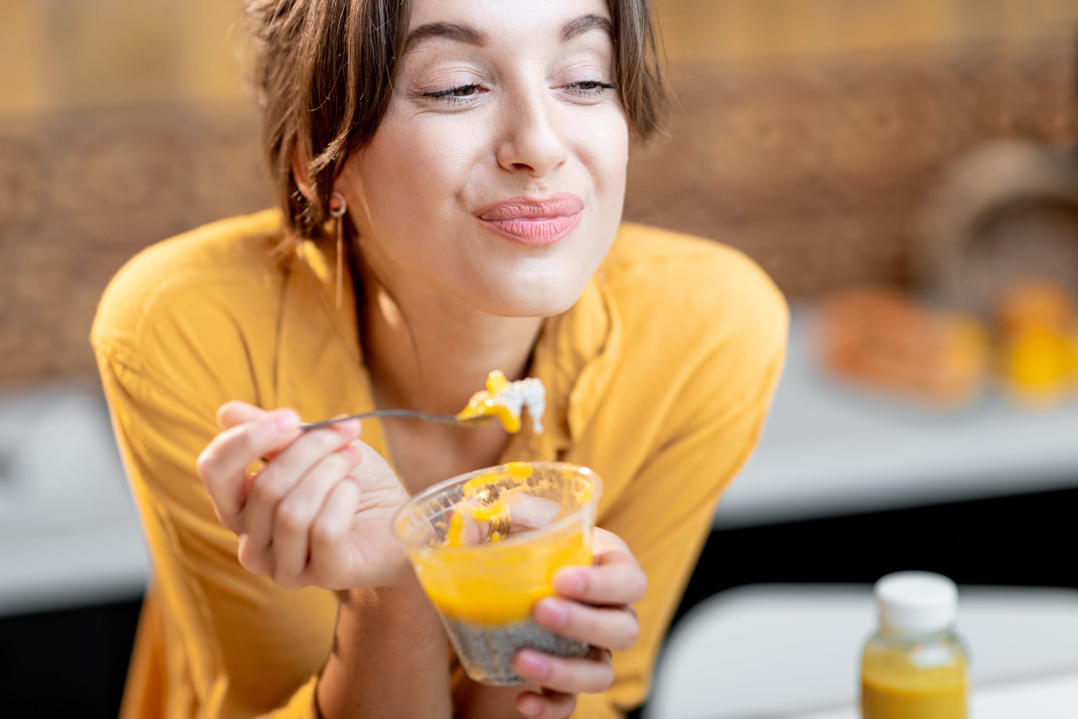 Woman wearing yellow top and eating chia pudding in kitchen.