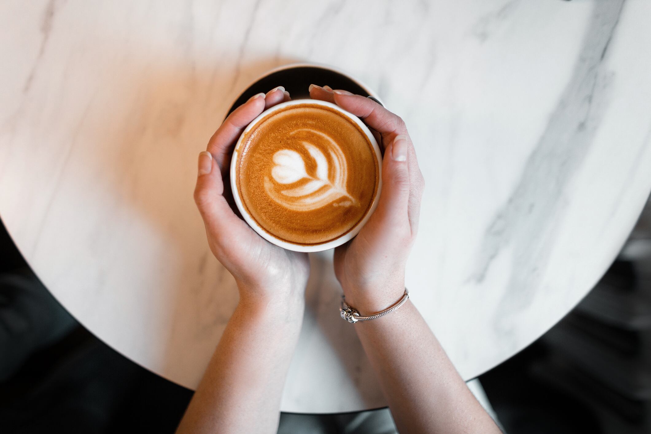 Female hands holding a cup of hot coffee with foam over a wooden table. View from above. Great morning.