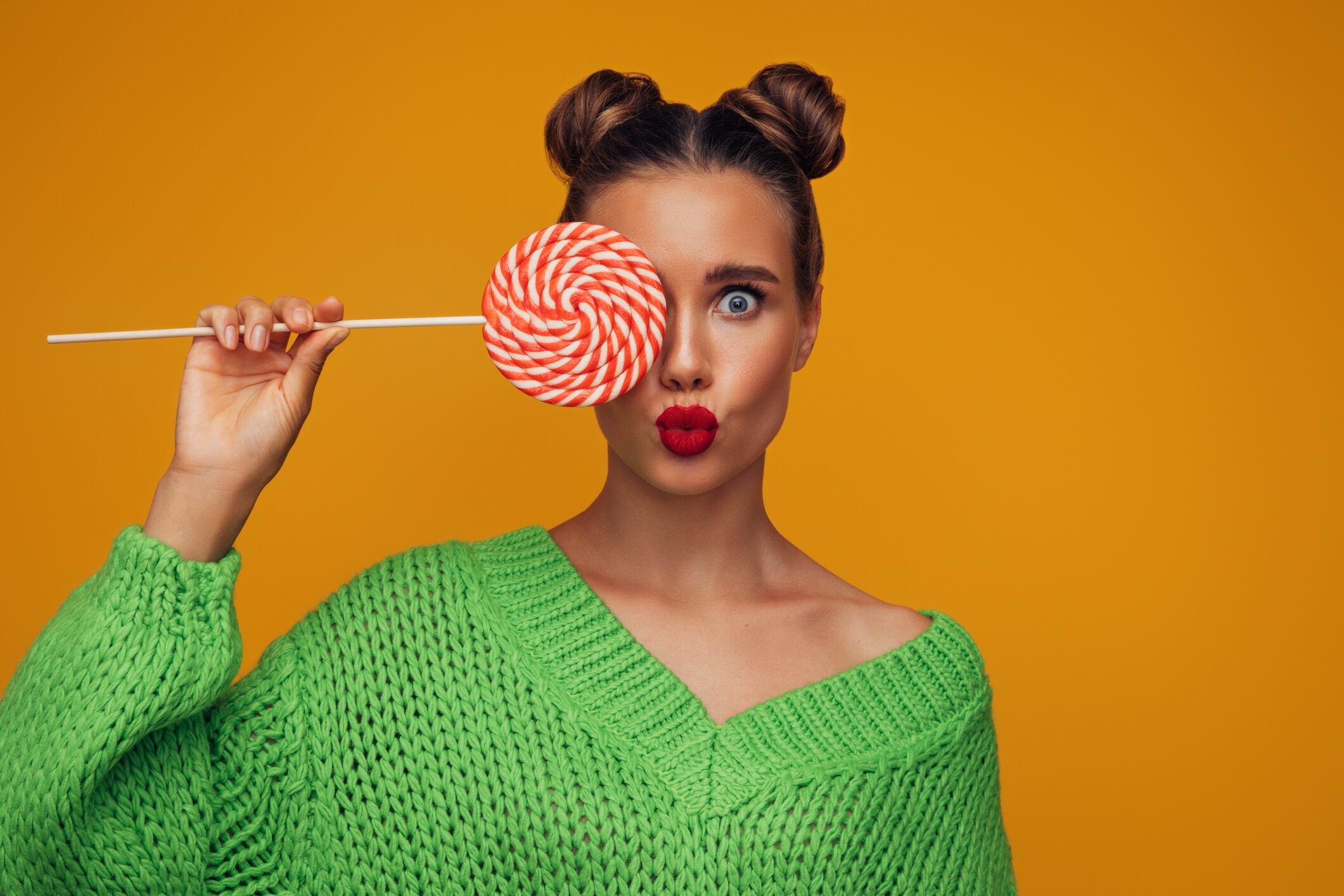 Woman wearing green knit jumper, wearing red lipstick, and holding lollipop over one eye.