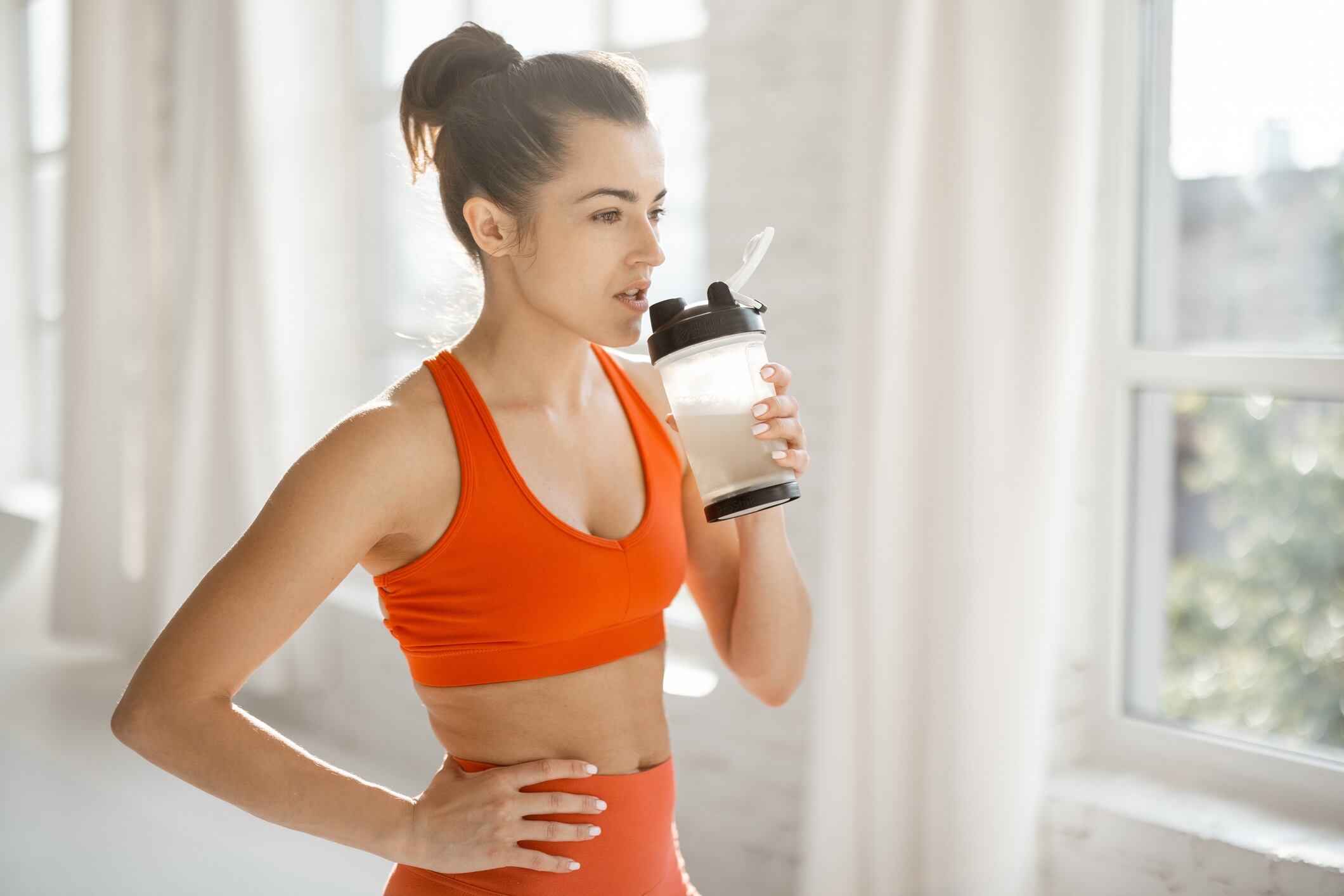 Athletic woman in bright orange sportswear drinking protein beverage from a shaker, standing indoors.
