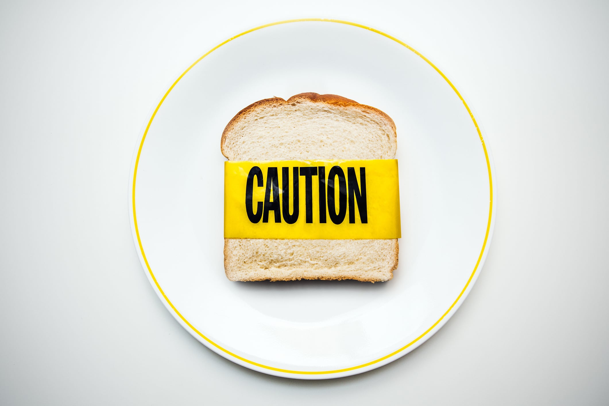A slice of white bread rests on a plate on a white table, yellow caution tape wrapped around it.  Conceptual image representing gluten or wheat allergies.