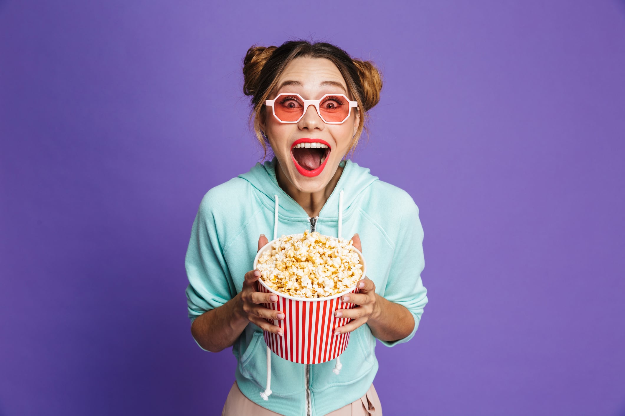 Woman wearing sunglasses, holding bucket of popcorn. Isolated on violet background.