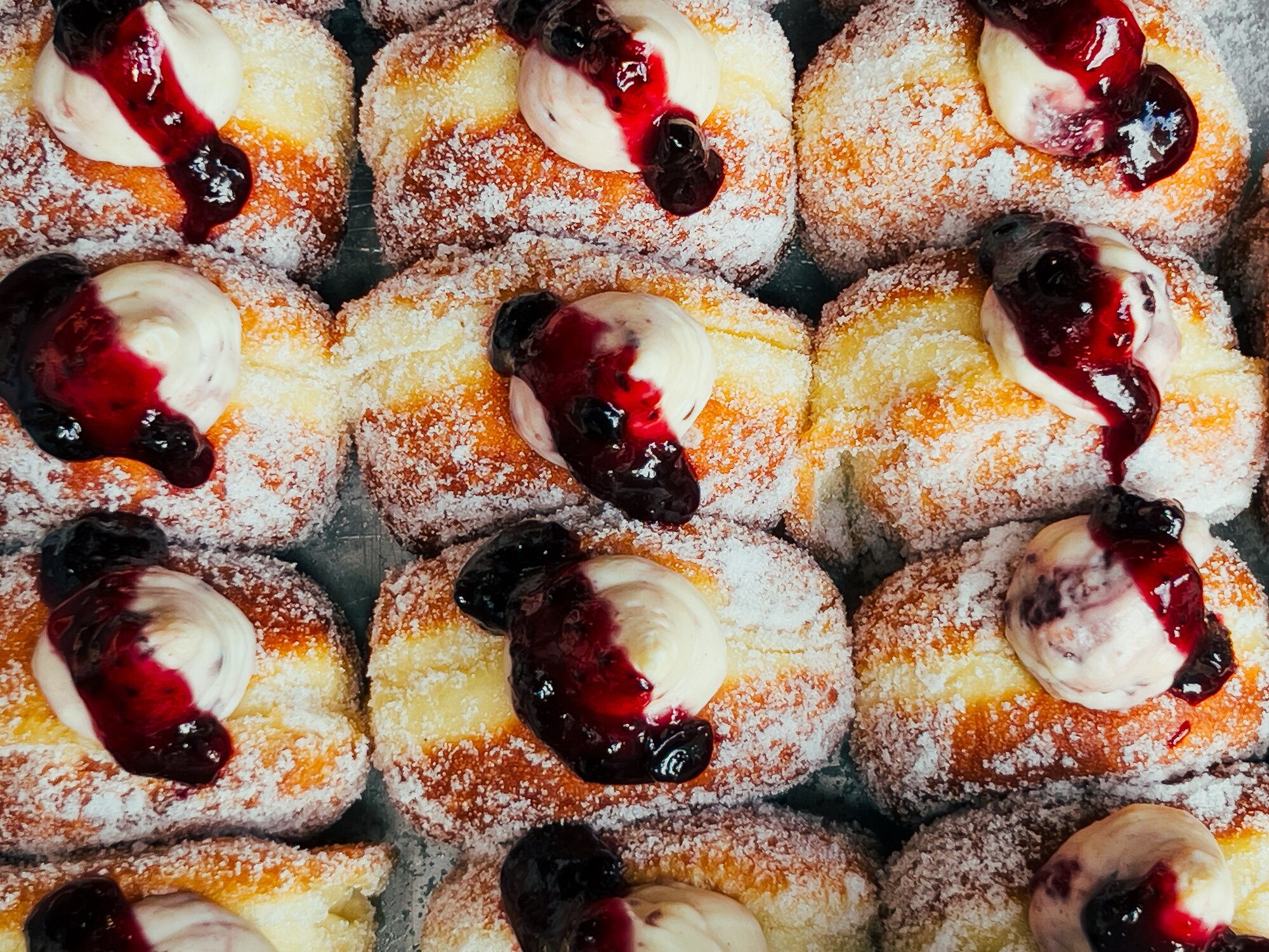 Color image depicting an overhead view of freshly fried doughnuts with different fillings including vanilla cream and raspberry jam for sale at a food market.
