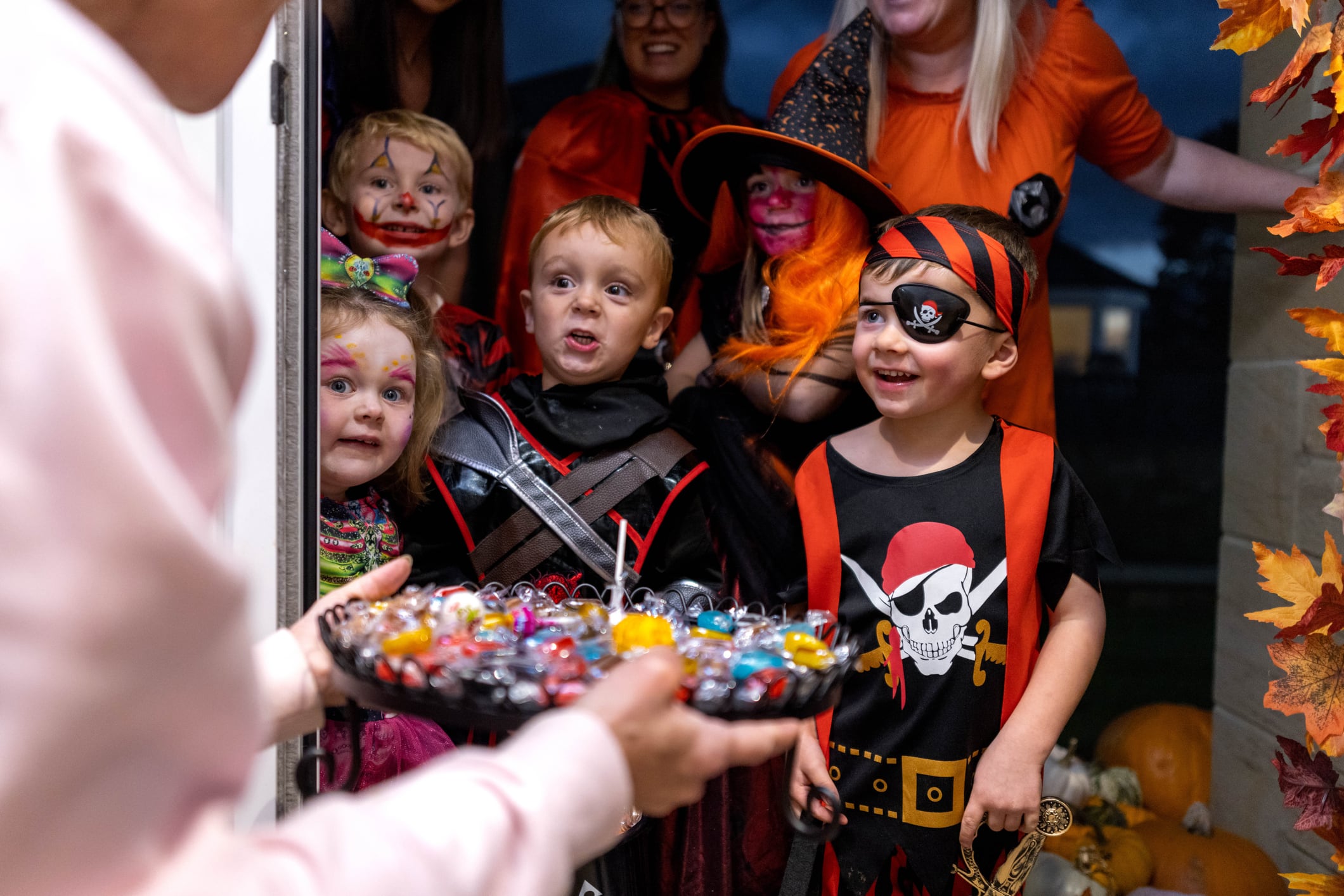 A small group of children trick or treating with adults standing behind them. Everybody is dressed up smiling and having a good time.
