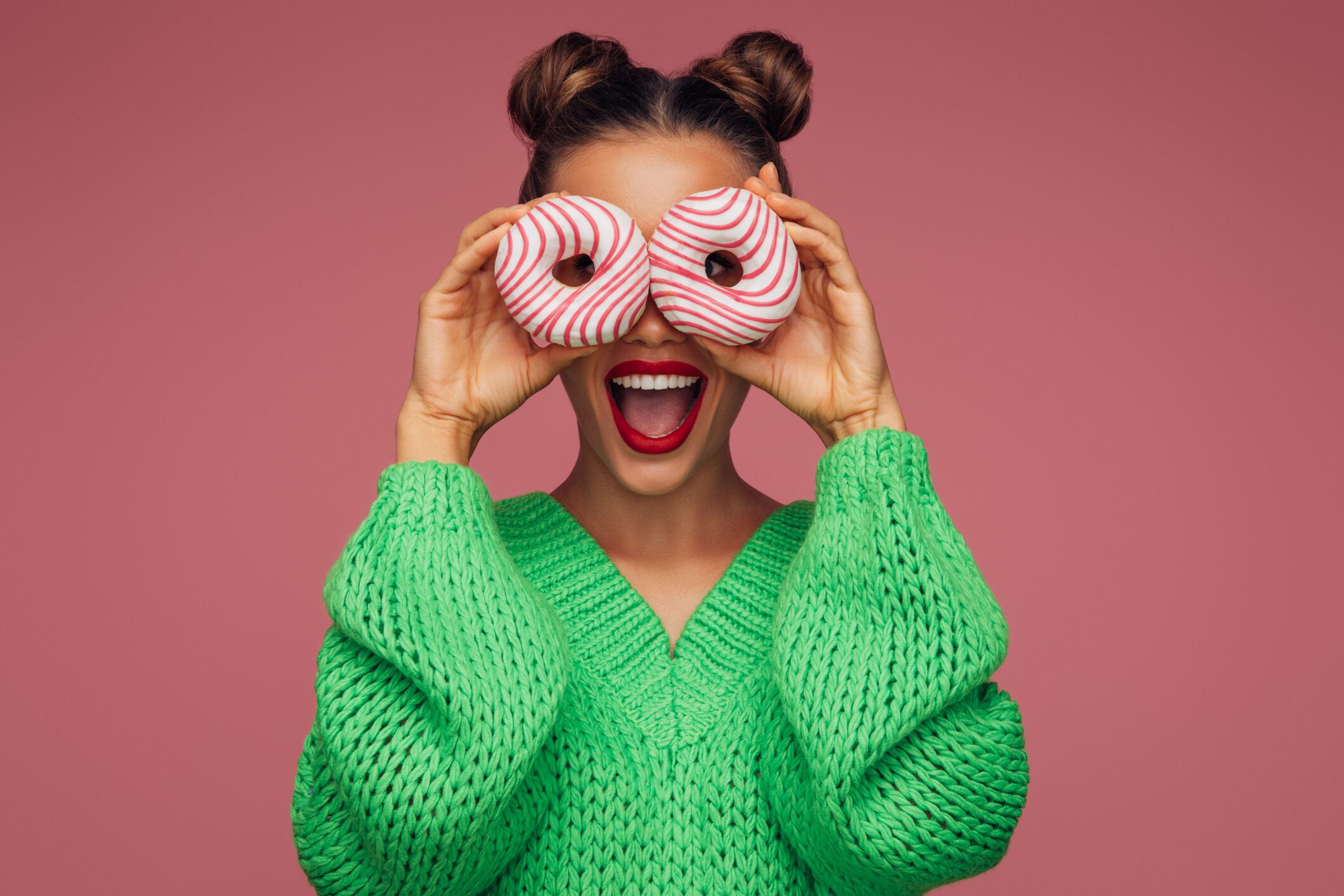 Woman holding doughnuts over eyes. Green woolen jumper and pink background.