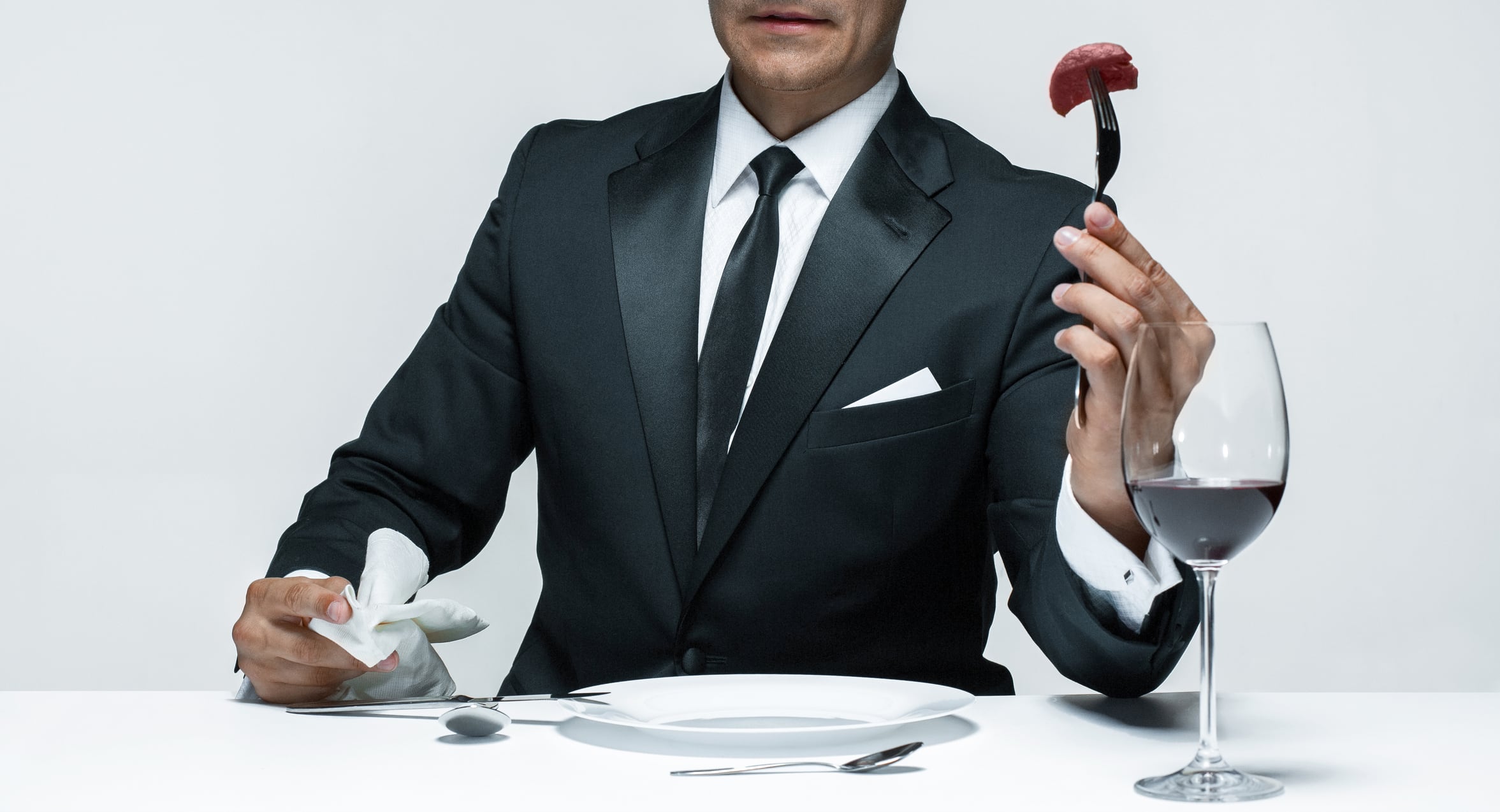 Man sat at dining table, wearing suit. He's holding a fork with a piece of raw meat on.