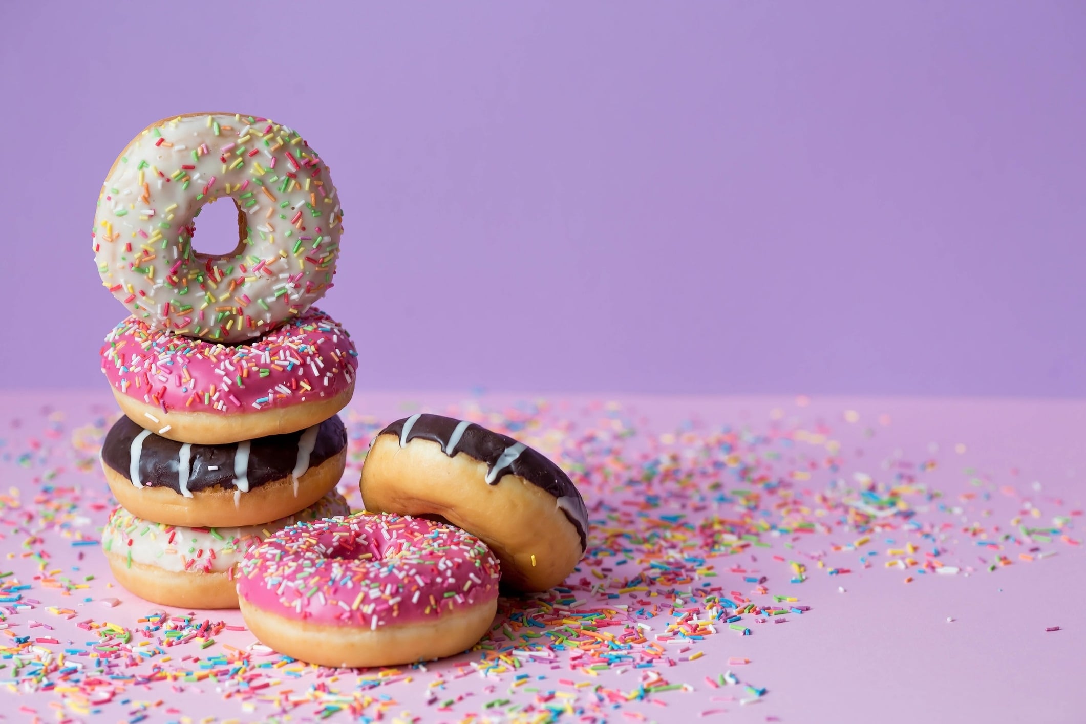 Pink, chocolate and white doughnuts, with multicolored sprinkles on a purple background.