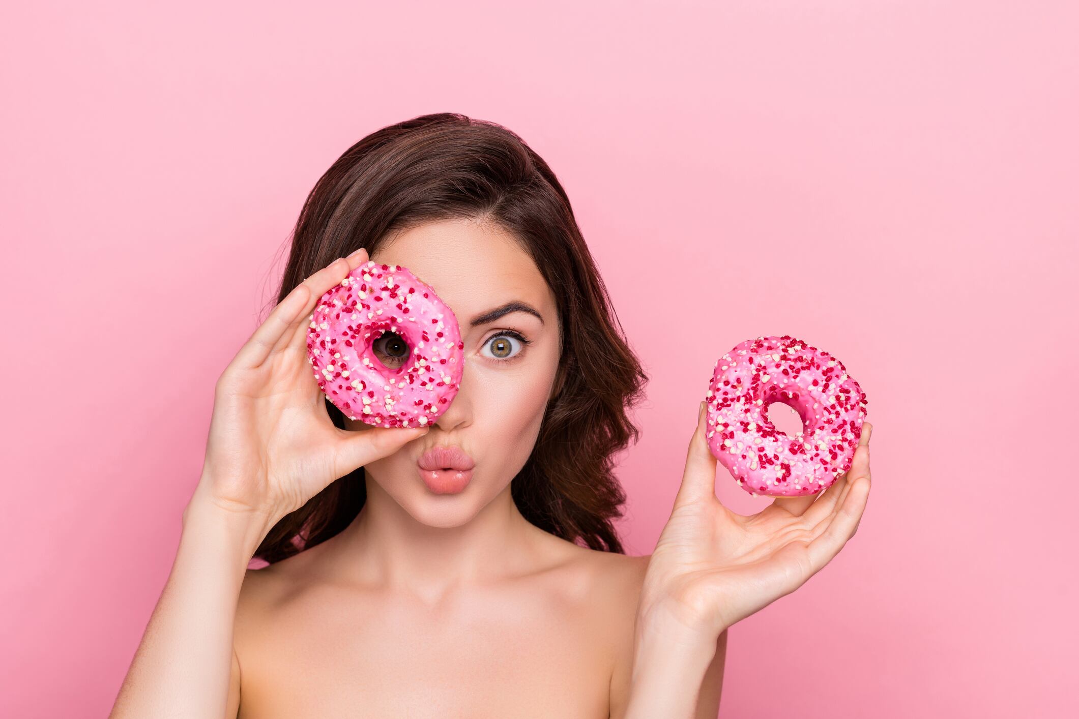 Woman holding two doughnuts - one over eye. Pink background.