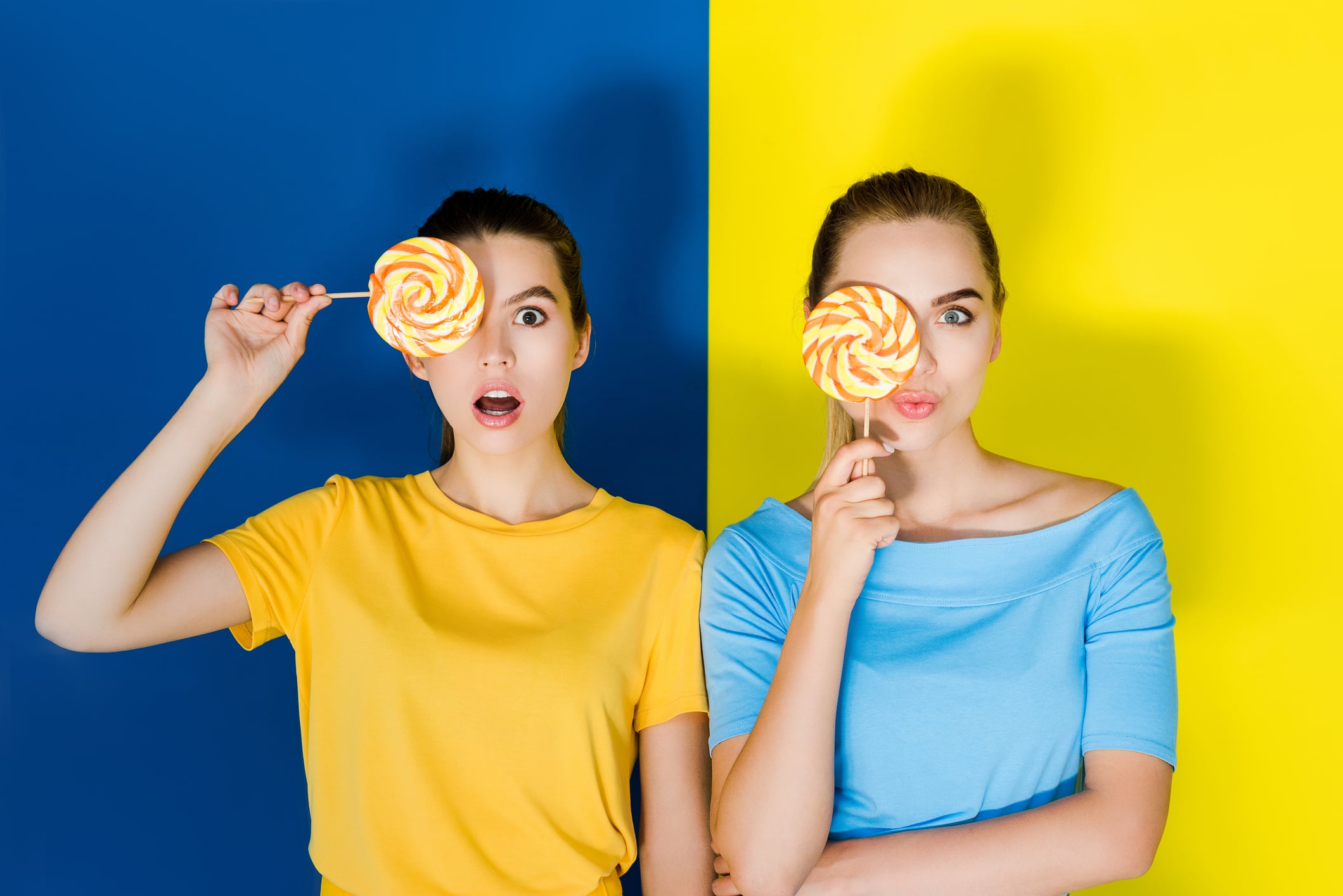 Two women holding lollipops on blue and yellow backgrounds.
