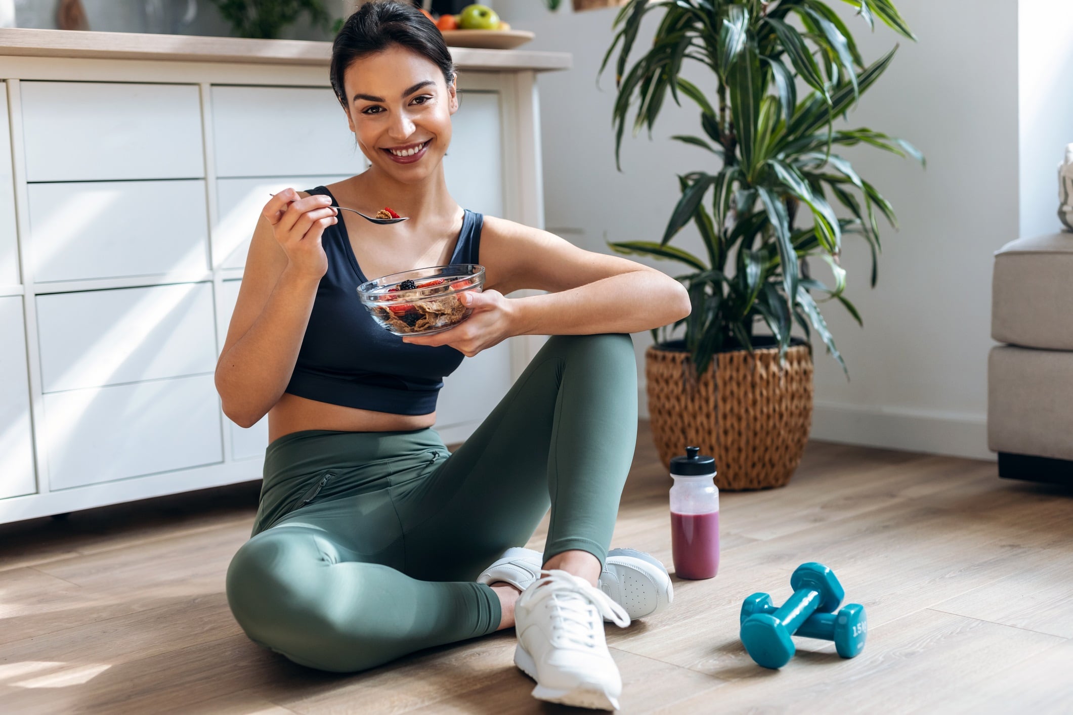 Shot of athletic woman eating a healthy bowl of muesli with fruit sitting on floor in the kitchen at home