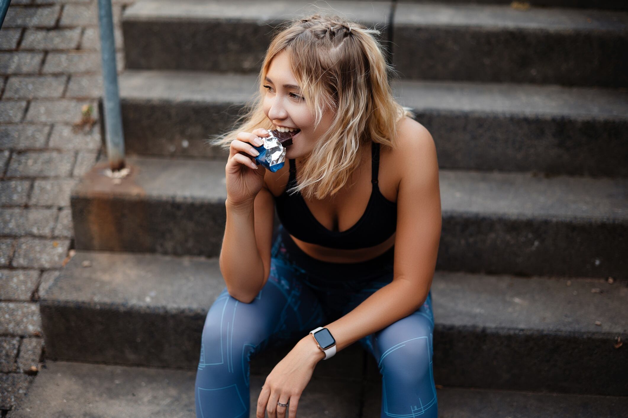 Woman with blonde hair, wearing sportswear, and sitting on steps, while eating fitness bar.