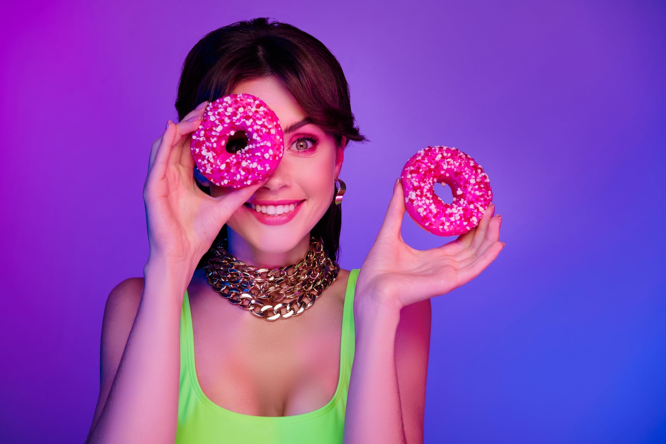 Woman holding two doughnuts - one covering right eye. Purple and pink background.